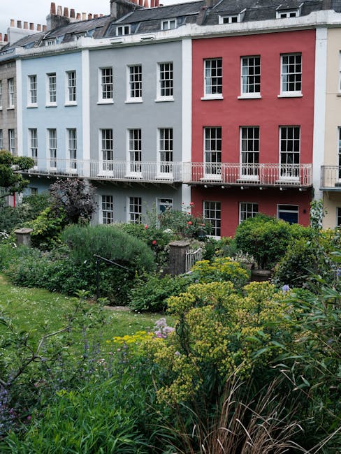 A multi-story terraced building painted in pastel shades of blue and pink, with white-framed windows and a narrow balcony extending along the upper floors. In the foreground, there is a lush, well-mai